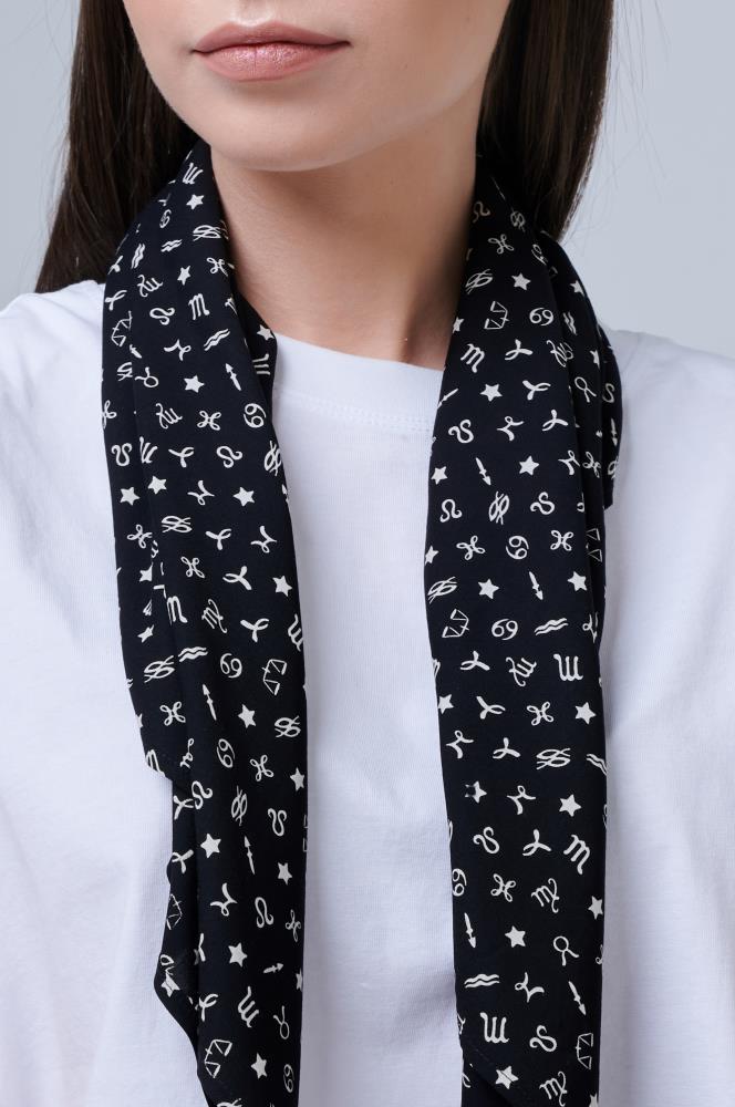 A close-up portrait of a woman from the lips to the chest, showcasing a dark scarf. The woman is wearing a simple white T-shirt, and draped around her neck is a black scarf decorated with small white zodiac sign symbols and stars. The ends of the scarf hang freely in front, creating a contrast with the light-colored garment and the neutral background.
