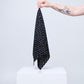 minimalist photo against a light background, showing a tattooed arm holding a black scarf by its corner. The scarf is decorated with small white zodiac sign symbols and stars. The accessory hangs above a white cube-shaped pedestal, with the composition highlighting the product's pattern and lightness.