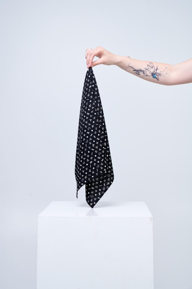 minimalist photo against a light background, showing a tattooed arm holding a black scarf by its corner. The scarf is decorated with small white zodiac sign symbols and stars. The accessory hangs above a white cube-shaped pedestal, with the composition highlighting the product's pattern and lightness.